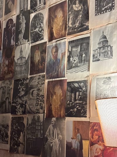 The image displays an interior wall in Varbak, Bulgaria, completely covered with a dense, overlapping arrangement of printed reproductions. These prints, varying in size, orientation, and color, predominantly feature classical artworks, architectural studies, and portraits. Visible subjects include black and white reproductions of a classical statue, a queen in elaborate attire, St. Paul's Cathedral, a domed building, and a Dutch canal house street scene. Other prints show a self-portrait of an artist (resembling Van Gogh), an interior scene with figures and a checkered floor (reminiscent of Vermeer), a seated old man in a robe, and a woman reading a letter. Some prints are sepia-toned or full-color, while others are in black and white. Many prints exhibit creasing, curled edges, and discoloration along their borders, indicating they are affixed directly to the wall. The scene is illuminated by warm, indirect light. A segment of a light-colored, mesh-textured lampshade is partially visible on the right side of the frame.