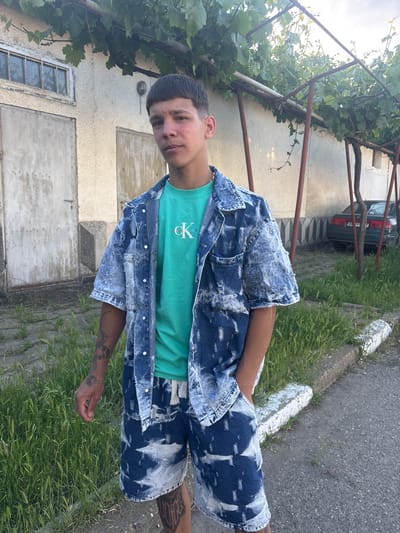 A young man stands on the edge of a road in Hayredin, Bulgaria. He wears a blue and white tie-dye denim jacket and matching shorts, and a light green t-shirt. Tattoos are visible on his arms and legs. Behind him is a light-colored building with peeling paint and weathered doors and windows, draped with grape vines. A dark car is partially visible behind the grape vines. The road runs along the edge of a grassy area.