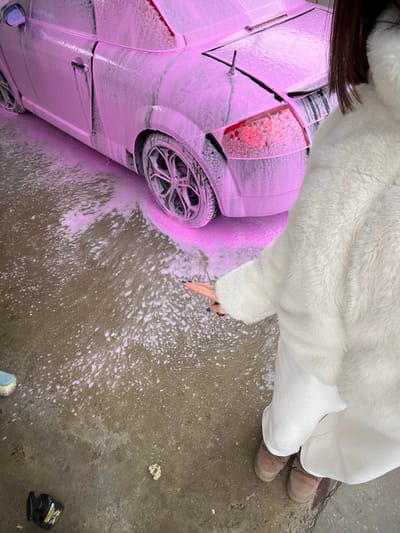 A two-door coupe, likely an Audi TT, is positioned on a wet, gray concrete surface in Bryastovo, Bulgaria. The vehicle's passenger side, rear bumper, and rear wheel are heavily coated in thick, vibrant pink car wash foam. The foam also covers the rear taillight, through which a red glow is visible. The rear wheel features a multi-spoke dark metallic rim. A person, partially visible in the foreground from the chest down, wears a white fluffy jacket, white trousers, and brown lace-up boots. Their right arm is extended towards the foamy ground, revealing fingers with blue nail polish. Dark brown hair is visible at the top right of the frame. The concrete floor is extensively wet and splattered with pink foam. In the lower-left foreground, a blue-handled brush and a small black container with yellow labeling are visible on the ground. A crumpled, light-colored object lies near the person's feet. The scene depicts a car being washed.