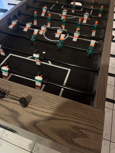 A foosball table is visible from an oblique top-down perspective. The black playing surface features white lines delineating the field and goals. Players, alternately colored white with green or orange accents, are mounted on black rods. The wooden table frame has a textured greyish-brown finish. The rods extend horizontally across the table. A portion of the floor with grey and white tiles is visible in the background.