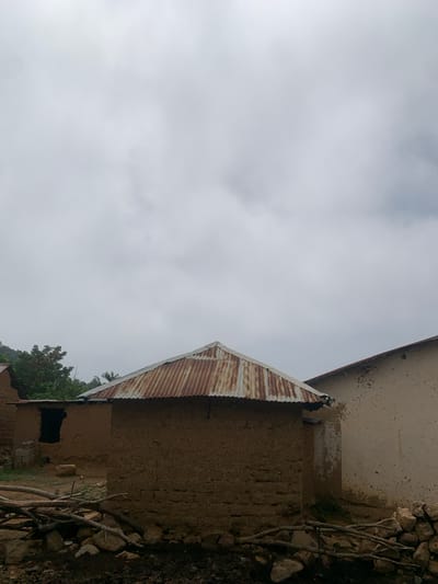 The image depicts a rural setting in Zhwarki Mile 10, Nigeria, under an overcast sky. In the foreground, a pile of wood and stones forms a rough barrier. Two mud-brick structures with corrugated metal roofs are visible. The building on the left has a dark, open doorway and appears to be under construction or in disrepair, with some stacked concrete blocks nearby. The building on the right is plastered with a light-colored material, showing signs of wear and tear. Green foliage, including trees, is present in the background, suggesting a vegetated environment. The overall impression is of a humble, possibly underdeveloped, rural community.