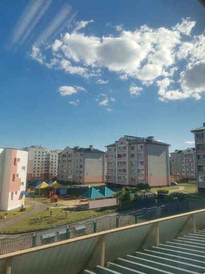 This media file captures a daytime scene in Ashmyany, Belarus, under a bright blue sky dotted with fluffy white clouds and streaky cirrus formations. The view looks out over a residential area composed of multi-story apartment buildings with light-colored facades, some accented with pink and green. Several of these buildings have visible numbers, including "95" and "93."
In the foreground, a children's playground is visible, featuring yellow and blue canopies, a red and yellow play structure, and a swingset. Green grass covers the playground area and the surrounding grounds, with some small trees and bushes interspersed. A blue metal fence with decorative elements encloses a portion of the playground area. Beyond the playground, there are more apartment buildings, a paved walkway, and parked vehicles, including a dark-colored SUV and a smaller dark car. The sky suggests fair weather, likely late morning or early afternoon due to the bright sunlight and the clear visibility. The overall impression is of a modern, well-maintained urban or suburban residential environment.