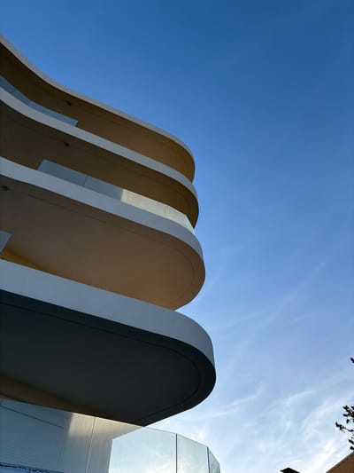 The image depicts an architectural structure with a series of curved, parallel fins or arches. The structures are white with tan-colored under surfaces and are set against a clear blue sky. The fins appear to be part of a building, and their repeating, rounded shapes create a sense of rhythm and movement. A section of the facade of the building with a combination of smooth and reflective material is seen in the right of the frame. A bit of foliage is present in the upper right corner. Considering the architectural style and the lack of information about the location, it is not possible to provide a more detailed description or confirm the location in the city of Municipal Unit of Elliniko, Greece.