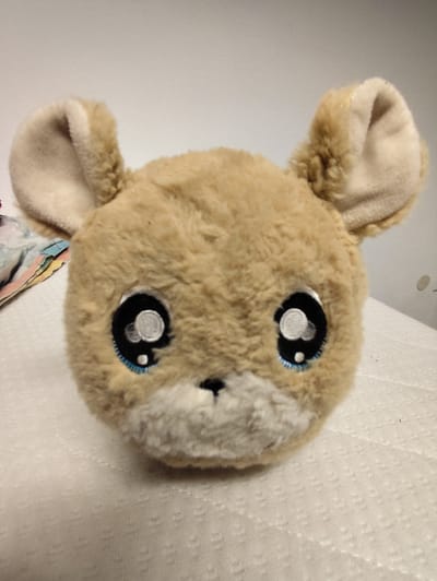 The image shows a close-up, head-on view of a tan-colored, fuzzy stuffed animal with large, embroidered black eyes that have white highlights, giving it a kawaii or anime-inspired appearance. The stuffed animal has two floppy ears, a lighter-colored fuzzy snout with a small black nose, and a hint of blue stitching around the lower edge of its eyes. It is positioned centrally in the frame, resting on a textured white surface that appears to be a mattress or a similar fabric. To the left of the stuffed animal, a small portion of a colorful fabric, possibly a blanket or quilt, is visible. The background is a plain, light-colored wall. There is no indication of the city of Schwäbisch Gmünd, Germany, in the image.