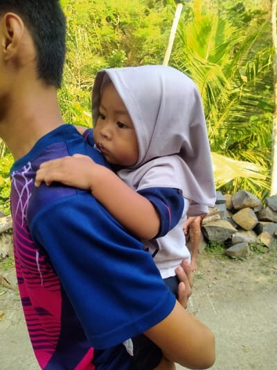 An outdoor scene in Jetis, Indonesia, depicts a partial view of a male figure carrying a young child. The male, positioned with his back and left side towards the viewer, wears a dark blue short-sleeved shirt with pink and magenta abstract patterns on the upper back and right shoulder. The child, held on the male's left arm, is seen in profile, looking towards the left side of the frame. The child wears a light grey hijab covering their head and a white and dark blue short-sleeved top. Their right arm is wrapped around the male figure's neck. The background is comprised of dense green foliage, including discernible palm fronds, under natural daylight. A pile of irregular grey and brown rocks is visible in the lower right background, situated near a light-colored ground surface.