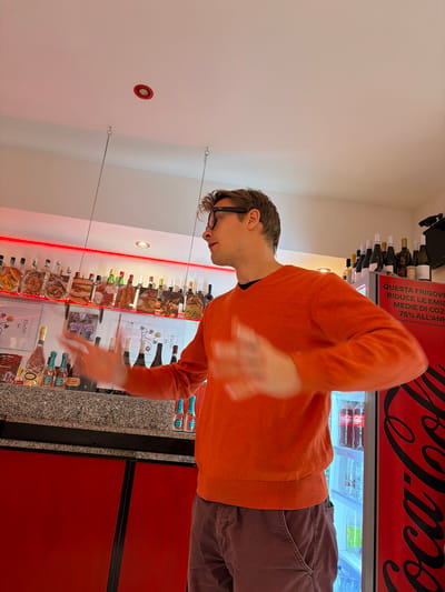 The image shows a man with glasses and an orange sweater standing in a shop, likely in Ragusa, Italy, based on the Italian writing on a cooler. He is facing a display shelf stocked with bottles of wine, and the cooler behind him has a Coca-Cola advertisement. The shelf seems to display advertisements and pictures of food, suggesting it might be a delicatessen or a specialty food store. The man's hands are slightly blurred, indicating movement. The lighting is bright, and the scene appears to be indoors.
