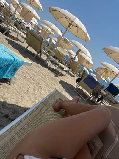 The image depicts a beach scene in Finale Ligure, Italy. In the foreground, a person's legs are visible, resting on a beige sun lounger. Their feet are bare, with painted toenails. The sun lounger has a light-colored frame and a woven beige surface. A blue towel with yellow detailing is draped over another sun lounger in the mid-ground, to the left of the frame. The beach is covered in light-colored sand. Rows of empty sun loungers and closed white beach umbrellas are arranged on the sand, suggesting it is either early morning or late afternoon, or a less crowded part of the day. In the background, the blue sea is visible under a clear blue sky. The overall impression is one of a tranquil beach setting.