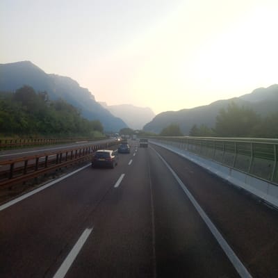 The image depicts a highway in the vicinity of Rivalta, Italy, characterized by a hazy, overcast sky and a backdrop of mountainous terrain. The highway is paved with asphalt and marked with white lane lines. Several vehicles are visible, including a dark-colored SUV in the foreground with a European license plate, a dark blue hatchback behind it, and other cars and trucks further ahead. A metal guardrail runs along the right side of the highway, separating it from a sound barrier made of concrete and glass. Dense green foliage and trees line the left side of the highway, climbing the slopes of the mountains. The lighting suggests it is either early morning or late evening, with soft, diffused light casting a muted glow over the landscape. The atmosphere appears calm and quiet, with no visible individuals outside of the vehicles.