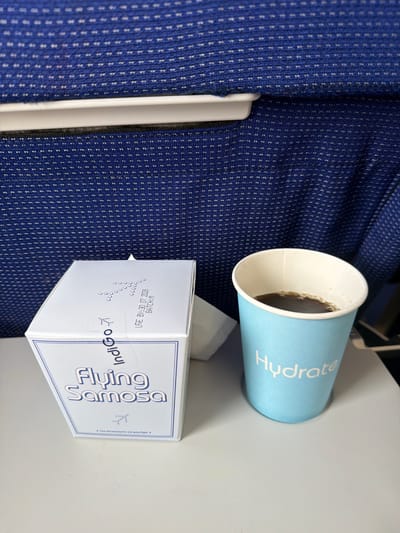 An overhead shot shows a blue Indigo Airlines branded box labeled "Flying Samosa" next to a light blue cup with the word "Hydrate" printed on it, containing a dark beverage. The items are on a light-colored tray table, likely in an airplane cabin, with the blue, patterned upholstery of airplane seats visible in the background. There are no people visible in the image. The scene is a close-up of the tray table, with the focus on the food and drink items. The specific location in Mohan, India, is not discernible from the image.