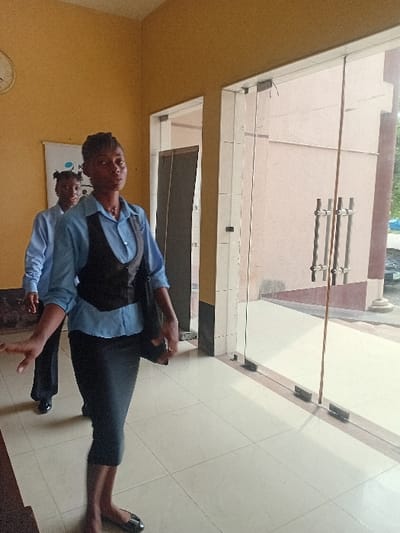 The image portrays an indoor scene, likely a building lobby or hallway, in the city of Ganme, Nigeria. Two African women are in motion, walking from left to right. The foreground subject is an adult female with dark skin and braided hair, wearing a light blue collared shirt, a dark vest, a dark pencil skirt, and dark flat shoes. She carries a dark shoulder bag on her left side and has her right arm extended forward, looking directly at the camera. Behind her, a second female, partially obscured, wears a similar light blue collared shirt and dark trousers, with her hair styled in two dark puffs. She is looking slightly to the left of the camera. The interior features light mustard yellow walls and a floor tiled with light beige square tiles. A round white wall clock, displaying approximately 10:50, is mounted on the upper left wall, above a white banner with a partial blue logo. On the right side of the frame, large transparent glass doors with vertical silver handles offer a view to the exterior, revealing a light brown building facade, pavement, and green foliage in the background under daylight. A dark, open doorway is visible on the far left of the image.