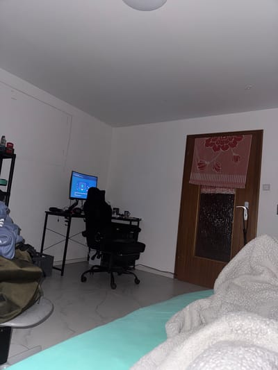 The image depicts a room that appears to be a bedroom or a multipurpose living space. The room is sparsely furnished and somewhat cluttered. On the left side of the image, there is a black metal shelf unit with various items on it, including bottles and what appears to be clothing. Next to the shelf, a large olive green bag is placed on a round object, possibly a stool or a small table.
In the center of the room, a black desk with a metal frame supports a computer monitor, a keyboard, a mouse, and other accessories such as headphones and a mug. A black gaming chair is positioned in front of the desk. The monitor displays a blue screen with some graphical elements, suggesting it is either a game or an application.
To the right, a wooden door is visible. A pink towel with a red floral pattern is draped over the top half of the door. The lower half of the door has a frosted glass panel. A silver door handle is present.
The floor is tiled with a light-colored marble pattern. In the foreground, a portion of a bed or a mattress covered with a light blue sheet and a fluffy white blanket is visible.
The lighting in the room is dim, likely artificial, with a ceiling light fixture partially visible at the top of the frame. The overall impression is of a lived-in space, possibly a student's room or a casual living area.
There are no people visible in the image. The scene does not provide any specific clues that would allow for the determination of its location within the city of Leverkusen, Germany.