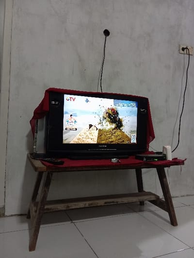 The image depicts an indoor scene in Bulukandang, Indonesia. A black CRT television, partially draped with a red fabric, rests on a rustic, dark brown wooden table featuring a lower shelf. The television screen displays content from "GTV," showing an animated or graphical scene with a character on the left, a large explosion or volcanic eruption in the center, and a split-screen effect with a textured background. A black remote control, a black electronic set-top box with an illuminated green LED, and a white cylindrical object (possibly a candle) are positioned on the table to the right of the television. The background consists of a light grey, textured wall. A black electrical cord extends from a white wall outlet located high on the right side of the wall, leading downwards out of frame. Another thin black cable, possibly an antenna wire, protrudes from a small hole in the wall above the television. The floor is covered with light-colored tiles. No individuals are visible in the scene.