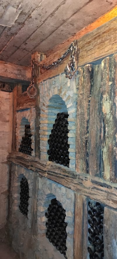 The image depicts a cellar in Voskevaz, Armenia, with stone alcoves built into a wall, each filled with stacked dark glass bottles, likely wine bottles. Heavy chains hang from wooden beams in the ceiling, with one chain connecting to a metal ring. The wall is constructed with rough-hewn stone and brick, and the ceiling is made of wooden planks. The overall impression is of a rustic and historical wine cellar.