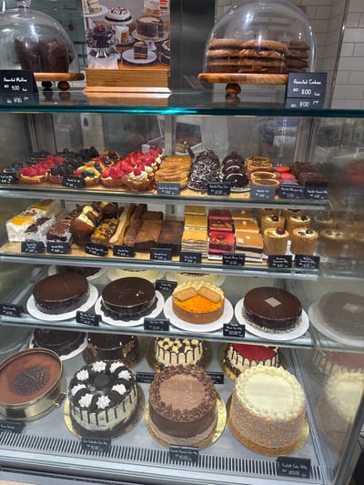 The image displays a refrigerated display case filled with various baked goods, including cakes, pastries, muffins, and cookies. The display case appears to be in a bakery or patisserie. The lighting is bright, highlighting the items in the case. Price tags are visible for some of the items, indicating prices in AED (United Arab Emirates Dirham). The environment suggests a commercial food establishment.