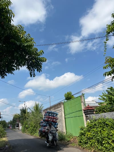 An outdoor, eye-level, medium shot shows a man riding a motorcycle laden with a large stack of balloons on a street in Jati Alun Alun, East Java. The man, wearing a light brown jacket and a helmet, is positioned in the lower-right quadrant of the frame. He is driving on an asphalt road, bordered by lush green vegetation on both sides. To the right of the road, a tall, gray concrete wall with a large, green metal gate is visible, partially covered by dense green foliage. Beyond the gate, a traditional red-roofed structure peeks out. In the background, a few houses and palm trees can be seen. The sky is a vibrant blue, dotted with fluffy white clouds. Power lines stretch across the upper portion of the image. The overall scene depicts a common sight in the region, with the motorcycle acting as a mode of transport and delivery for goods.