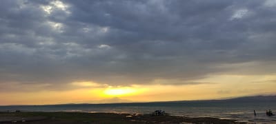 A wide shot captures the sunset over a large body of water, likely a lake or sea, with mountains visible in the distance. The sky is filled with dramatic clouds, with a bright patch of sunlight breaking through above the horizon, casting a warm yellow and orange glow. In the foreground, a dark shoreline is visible, with some debris and equipment scattered on it. To the right, a small boat with two people in it is visible on the water. The overall atmosphere is one of a peaceful evening at the end of the day. The image is believed to have been taken in the city of Chigdalik, Tajikistan.