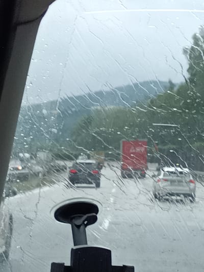 This media file captures a view from inside a vehicle, looking through a rain-streaked windshield. The scene is set on a highway during what appears to be a rainy day, with water droplets and rivulets distorting the view of the road and surrounding landscape. In the distance, a blurred, hilly terrain covered in green foliage suggests a rural or semi-rural location.
Several vehicles are visible on the wet asphalt road. Directly ahead, a dark-colored car with its taillights illuminated is driving in the same direction. To the right, a white car is also moving forward. Further ahead and to the left, a large red truck is a prominent feature on the highway. The overall impression is of traffic moving through inclement weather.
A suction cup mount for a device is visible in the foreground, attached to the windshield, indicating the perspective is from a driver or passenger. The time of day is unclear due to the overcast and rainy conditions, but the general ambiance is subdued and possibly melancholic, typical of driving in the rain. No discernible text is visible in the image. The location context provided is Hvězdonice, Czechia, which is consistent with the European appearance of the vehicles and the landscape.