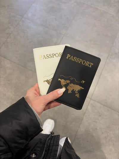 A person's right hand holds two passport covers, one black and one cream-colored, above a light grey tiled floor. The black cover is fully visible, featuring "PASSPORT" in gold lettering along with a gold world map design and a dashed airplane travel path. The cream-colored cover, partially obscured behind the black one, also displays "PASSPORT" in gold. The hand has manicured fingernails with white tips. The individual is wearing a black quilted jacket and a grey long-sleeved garment, with white athletic shoes visible on their right foot. The scene takes place in the city of Boldino, Russia.