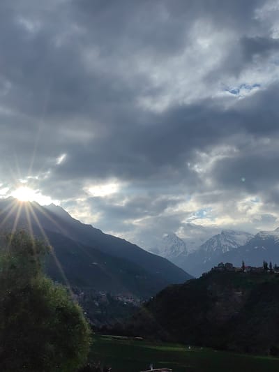The image captures a dramatic mountain landscape with the sun peeking through heavy, dark clouds. The sun's rays are visible, creating a luminous effect. In the foreground, there is a large, leafy tree on the left, partially obscuring the view. The middle ground features a hillside dotted with buildings, suggesting a settlement. Further in the background, majestic snow-capped mountains rise, their peaks contrasting with the cloudy sky. The overall scene evokes a sense of natural beauty and grandeur, with hints of human habitation within the vast mountainous terrain. This scene is located in the village of Kardang, India.
