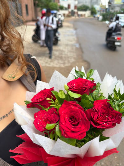 The image is a close-up shot of a bouquet of red roses, partially obscuring a person's shoulder and back. The roses are vibrant and dusted with gold glitter. The bouquet is wrapped in white and red paper, with some of the paper frayed at the edges. A tag is attached to the bouquet with the text "Earth..." visible. Below the bouquet, a tattoo is visible on the person's skin, but the text is too blurred to be legible. The background is out of focus, showing what appears to be a street scene with motorbikes and buildings, suggestive of a busy urban environment. The lighting suggests it is daytime. The overall impression is one of a gift being presented or received, with the focus on the beauty of the flowers.