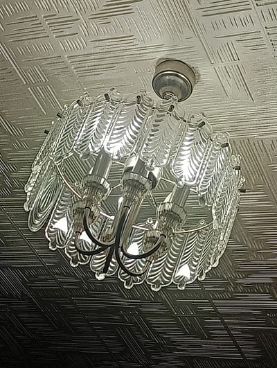 The image shows a chandelier hanging from a textured ceiling. The chandelier is made of clear glass elements shaped like feathers or leaves, with visible light bulbs illuminating the interior. The ceiling has a metallic or reflective finish with a raised, linear pattern. The lighting suggests an indoor setting, likely a room. The pattern on the ceiling transitions from a lighter, silver-toned area to a darker, greyish-black area, creating a gradient effect that also covers part of the chandelier. There is no visible text in the image.