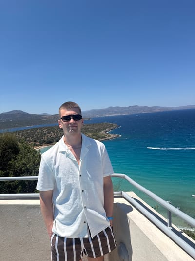 This is a portrait of a man standing on a balcony, overlooking a body of water. The man is wearing a white, short-sleeved button-down shirt and brown and white striped shorts. He has short hair and is wearing dark sunglasses. He appears to be in his late 20s or early 30s. The background shows a clear blue sky, mountains, and turquoise water with a boat in the distance. The setting appears to be a coastal area, likely a resort or vacation spot. The sunlight suggests it is daytime, and the clear sky indicates good weather. The man's relaxed posture and sunglasses hint at a leisurely atmosphere. The image's orientation is portrait, but the scene itself is captured from a high vantage point looking down and across. The location is identified as Agios Nikolaos Municipal Unit, Greece. There is no visible text in the image.