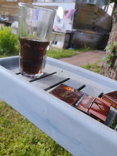 A glass of dark liquid, likely soda or iced tea, sits on a light blue plastic tray. Next to the glass are two open cigarette packs. The pack on the left is labeled "WINSTON" and the pack on the right is labeled "PAK TOPHA." The tray is placed on a surface covered with green grass. In the background, a rustic wooden building can be seen, along with some green foliage. The scene appears to be set outdoors, possibly in a backyard or garden area. There are no people visible in the image.