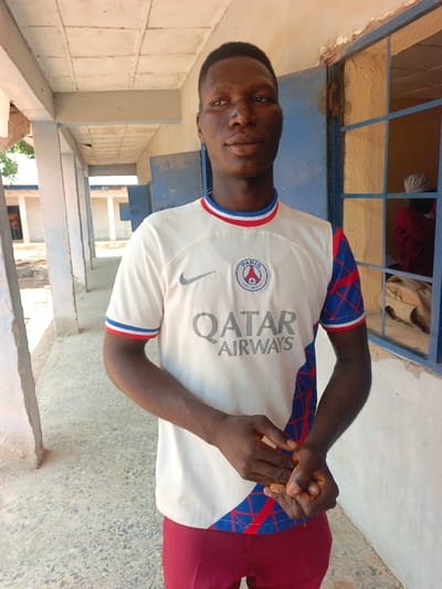 A male individual is standing under a covered concrete walkway in Mada, Nigeria. The subject, a dark-skinned male appearing to be in his late teens or early twenties, faces the camera with a neutral expression. He is wearing a white short-sleeved t-shirt featuring a "Paris Saint-Germain" crest, "QATAR AIRWAYS" text, and a Nike swoosh, complemented by blue and red trim on the collar and a blue patterned right sleeve. He wears dark red trousers and has his hands clasped together in front of his waist. The background on the left includes light-colored concrete pillars supporting a paneled ceiling, leading to an outdoor area with visible ground and a distant building with a blue door. To the subject's right, a window with a dark blue metal grid frame shows a partially obscured individual with a light-colored head covering and some fabric objects within a dimly lit interior. The scene is illuminated by ambient daylight.