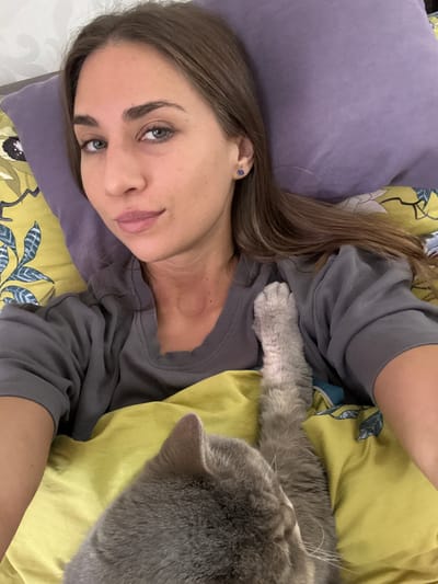 A woman is lying on a bed with a gray cat. The woman has light brown hair and is wearing a gray shirt. She is resting her head on a purple pillow. The bed has a yellow floral patterned sheet. The cat is lying on the sheet next to the woman, with its paw on her chest. The photo appears to have been taken from above. The location of the photo is unknown, and there is no specific indication that it was taken in Zanevka, Russia.