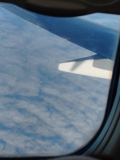 A view from an airplane window shows a wing against a backdrop of clouds. The wing is in the upper portion of the image, partially obscuring the sky. A white engine pod is visible below the wing. The clouds are white and fluffy, spread across a blue sky. The image is taken from a height, indicating an aerial perspective. The location is identified as Khrabrovo, Russia.