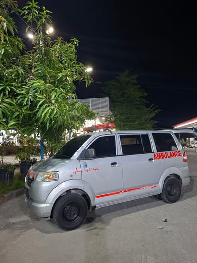 The image captures an ambulance parked at night, likely in Keuniree, Indonesia, given the context. The ambulance is silver with red lettering spelling out "AMBULANCE" on its side. It features a red emergency light bar on its roof and a red heartbeat line graphic with a heart emblem on the front fender. The vehicle is positioned on a paved surface, possibly a parking lot or road, with a large tree with green leaves to its left. In the background, there's a building with horizontal white louvers and some lighting. The scene is illuminated by artificial lights, creating shadows and highlighting the ambulance.