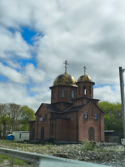 The image depicts an Orthodox church in Glebovskoye, Russia. The church is made of red brick, with two golden domes topped with Orthodox crosses. The sky is partly cloudy with patches of blue. The church is surrounded by trees and landscaping that appears unfinished. A portable toilet and construction materials are visible in the churchyard. The photograph appears to have been taken from a vehicle, as a guardrail is visible in the foreground.