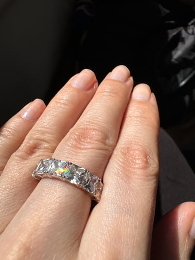 In the city of Vyatskiye Polyany, Russia, a close-up image captures the back of a person's left hand, illuminated by strong, direct light. A silver-toned eternity band, featuring multiple clear, square-cut stones, adorns the ring finger. One central stone prominently displays a vibrant rainbow spectrum due to light dispersion. The hand's skin exhibits natural texture, visible pores, and creases. The fingernails are short, neatly kept, and appear unpolished or with a clear coating. The background is uniformly dark and out-of-focus, suggesting an indistinct environment behind the hand, with minimal discernible features.