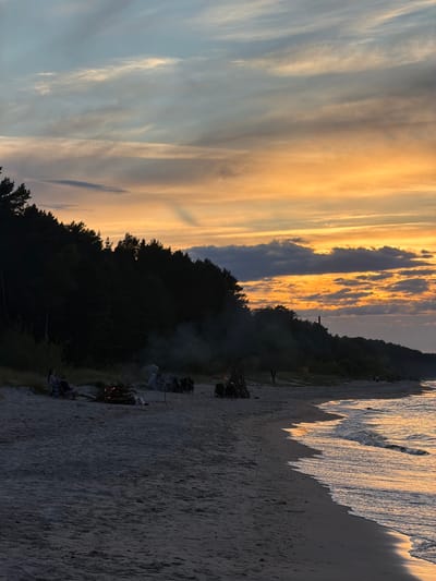 This media file captures a serene sunset scene on a beach in Plieņciems, Latvia. The sky is a canvas of warm hues, with streaks of golden orange and soft yellow clouds stretching across a pale blue expanse. Wispy clouds create a dramatic contrast against the darker, silhouetted treeline that forms the backdrop of the scene.

The foreground is dominated by a sandy beach, with the gentle waves of the sea lapping at the shore. The water reflects the golden light of the setting sun, creating a shimmering effect on the wet sand. Several bonfires are visible along the beach, with smoke rising into the air, indicating a gathering or event taking place.

In the mid-ground, scattered groups of people can be seen around the bonfires, some sitting in camp chairs, others gathered closer to the flames. The atmosphere appears relaxed and communal, with the soft glow of the fires adding a warm ambiance to the cooling evening air. The dark silhouettes of trees and hills frame the beach, adding depth and a sense of natural beauty to the location.

The time of day is clearly sunset, judging by the vibrant colors in the sky and the soft lighting. The weather appears calm, with no indication of wind or rain. The presence of beach, sea, bonfires, and people suggests a recreational gathering, possibly a camping trip or a beach party. No visible text is present in the image.