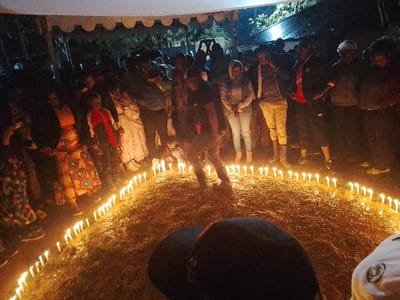 This nighttime image documents an outdoor gathering in Marenyo-Shianda ward, Kenya, occurring under a white canopy structure. Approximately 20-30 individuals, predominantly adults of varying ages, are present. A large circle of numerous lit white candles defines a central area on the dry grass ground. Within this candle circle, one individual in dark attire is observed bending forward or crouching. The surrounding crowd stands, observing the central activity; some individuals in the background are holding up mobile devices, possibly recording. Attire is casual, including patterned dresses, jeans, and jackets. The scene suggests a ceremonial event or vigil.