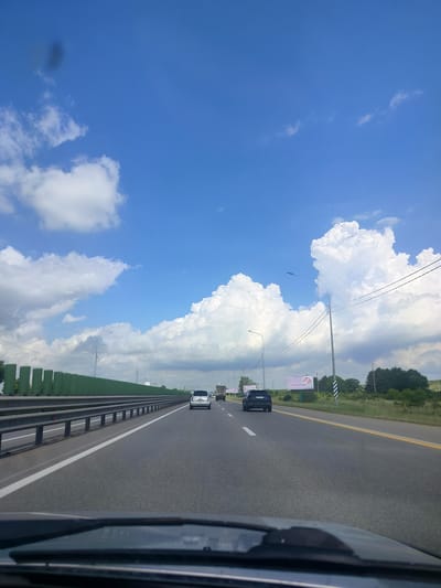 The image shows a highway with a blue sky filled with fluffy white clouds. In the foreground, the dashboard of a car is visible, indicating that the photo was taken from inside a moving vehicle. The highway has multiple lanes, and several cars are seen driving on it. On the left side of the road, there is a green sound barrier. On the right side, there are trees and a few billboards. The scene appears to be a sunny day in an open area, possibly outside of a city. The location is the city of Labinsk, Russia, on a highway.