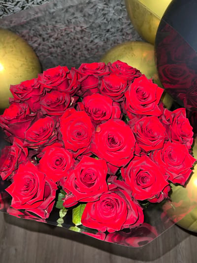 A bouquet of red roses is presented in a clear plastic wrap, with several gold and black balloons in the background. The roses are in full bloom, with deep red petals and visible textures. Some of the rose petals have slight imperfections or darker edges. The plastic wrap around the roses reflects light and the surrounding objects, creating a glossy effect. The balloons appear to be inflated and are positioned behind the floral arrangement. The overall setting suggests a celebratory occasion. There are no people or animals visible in the image. The background is partially obscured by the balloons and the bouquet, but it appears to be a textured surface, possibly a carpet or fabric. The lighting is somewhat dim, but the red of the roses is vibrant. There is no information in the image that would indicate the location in the city of Heorhiivka, Ukraine.