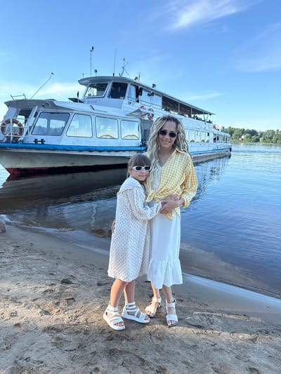 The image depicts two females, an adult and a child, standing on a sandy riverbank in Dumino, Russia. The adult female, with long blonde dreadlocks and dark oval sunglasses, wears a yellow and white checkered shirt over a white top, a white long skirt with a ruffled hem, and white flat sandals. She stands with her arms around the child. The child, with shoulder-length brown hair and white rectangular sunglasses, wears a white long-sleeved dress with small dark patterns and white platform sandals. She stands with her arms around the adult. Both individuals are smiling and looking towards the viewer.
Behind them, a large white passenger boat is docked in the calm, dark blue water. The boat features a blue stripe along its waterline and a red hull below it. Multiple windows are visible along its side, and an upper deck with railings shows several passengers. The inscription "Ярославский речной порт" (Yaroslavl River Port) is clearly visible in Cyrillic on the boat's forward superstructure. A white life buoy with partially visible Cyrillic text is attached to the boat's bow. The sky above is clear and bright blue, with minimal cloud cover. On the distant riverbank, a line of green trees and faint structures are visible. The foreground consists of light brown sand with numerous footprints. The scene is illuminated by bright, natural daylight, casting soft shadows on the sand.