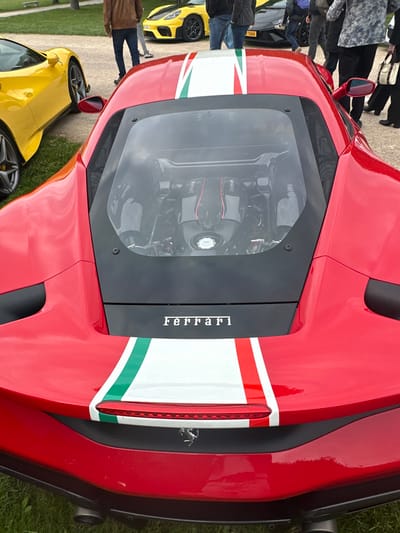 The image shows the rear end of a red Ferrari sports car, with a white and green racing stripe, parked in a grass lot. The car's engine is visible through a transparent cover. The car is parked near other cars, including a yellow sports car, and several people standing around. The location is near the Chateau de Chambord, a large French castle, located at 47.616 latitude, 1.518 longitude. The car is facing away from the camera. The camera is positioned to the right of the car, taking a rear angle shot. The Ferrari is the primary subject of the image, and it is in focus. The background is blurry, and the people are out of focus. There are no other details that can be gleaned from the image.