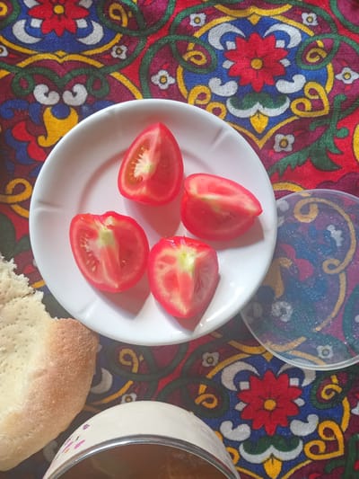 A close-up, top-down shot captures a white plate with four tomato wedges on a vibrantly patterned tablecloth. The tablecloth features a symmetrical design of red, blue, yellow, green, and white floral and swirling motifs. To the left of the plate, a piece of fluffy, light-brown bread rests on the tablecloth. To the right of the plate, a clear plastic lid with a colorful, abstract pattern is partially visible, also on the tablecloth. The image is set in the city of Kasansay, Uzbekistan.