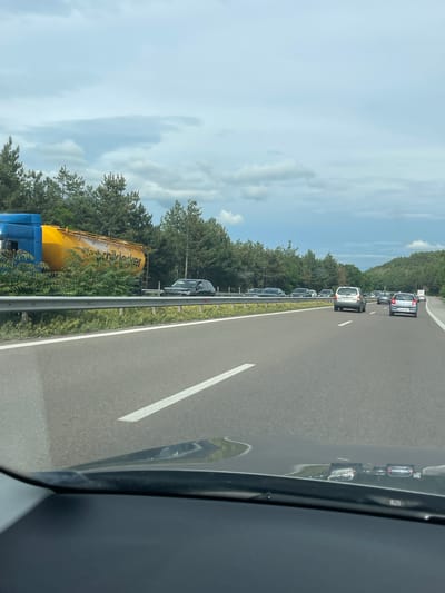 The image shows a highway with several cars driving, indicating traffic. On the left side of the highway, a large yellow and blue truck trailer is tipped on its side. The truck appears to be an industrial vehicle, possibly a cement or bulk material transporter, with the brand name "STRACKER" visible on its side. The trailer is partially obscured by foliage and a metal guardrail. The environment is characterized by a wooded area on the left side of the road, with a cloudy sky overhead. The road itself is paved and marked with white lines. The scene suggests a traffic incident, likely a rollover, has occurred. The location is identified as Gorno Varshilo, Bulgaria.