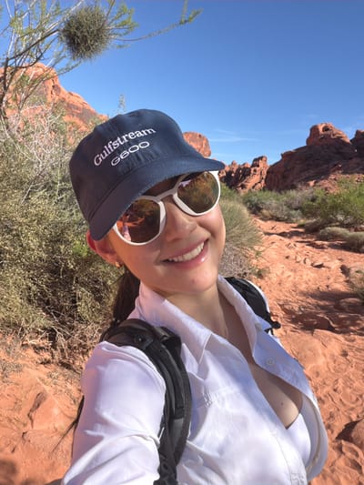 A medium-shot, slightly rotated selfie captures a woman in the landscape of Moapa Valley, United States. She wears a navy baseball cap, white-framed sunglasses, and a partially unbuttoned white shirt over a white top. A black backpack strap is visible on her right shoulder. The background consists of arid terrain with red rock formations, desert vegetation, and a clear blue sky. The woman is smiling, revealing her teeth. The reflection in her sunglasses shows someone in a similar attire.