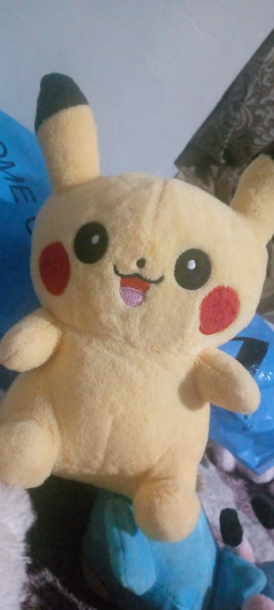 A stuffed toy resembling the Pokémon character Pikachu is the primary subject of this close-up image. The Pikachu toy is predominantly yellow with black tips on its ears and red circular markings on its cheeks. It has large black eyes with white pupils and a small, pink mouth with a tongue sticking out. The toy is positioned centrally in the frame, with its body filling most of the lower portion of the image.
Behind the toy, a bright blue bag is partially visible on the left side of the frame, with some dark text on it. To the right, a patterned fabric, possibly a blanket or upholstery, is visible. The background is a soft, light blue, likely a wall or the sky, though it is out of focus. The lighting appears to be artificial, casting a gentle glow on the toy.
The overall impression is that of a cute and cheerful toy. There are no people visible, and the setting appears to be indoors, possibly a bedroom or living area. The image seems to be taken in Marhamat, Uzbekistan, as indicated by the location context. There are no obvious cues about the time of day or weather. The toy is static, with no particular activity taking place, other than its display.