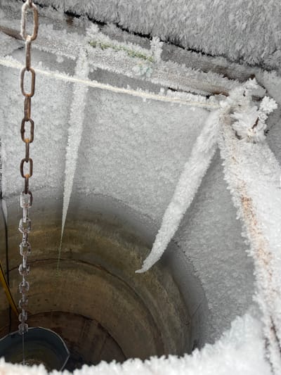 The image displays the interior of a water well in Карабоўскі сельскі Савет, Belarus, viewed from directly above, looking down into the shaft. The upper walls and internal structures of the well are uniformly coated in dense, white hoarfrost, forming crystalline, feathery textures and numerous elongated, downward-hanging frost formations. A rusted metal chain, partially encased in clear ice, descends vertically along the left side of the frame. Towards the bottom left, a metallic bucket is suspended by this chain, partially submerged in visible water at the well's base. Several ropes and cables, also encrusted with frost, are visible spanning across the upper interior, including a green-colored rope and a yellow cable. The visible lower section of the well walls consists of circular concrete or stone rings, showing evidence of water staining and dampness.