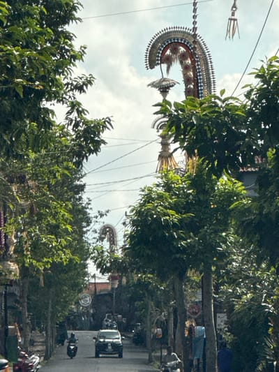 In Bangli, Indonesia, a street is lined with lush green trees, casting shadows on the asphalt road. Overhead, elaborate decorations, possibly for a festival, are suspended from wires, with one prominent, ornate structure featuring intricate patterns and hanging elements. A white car drives down the center of the road, with a person on a motorbike visible in front of it. A few other individuals are seen along the sides of the street. The sky above is a mix of white clouds and blue.