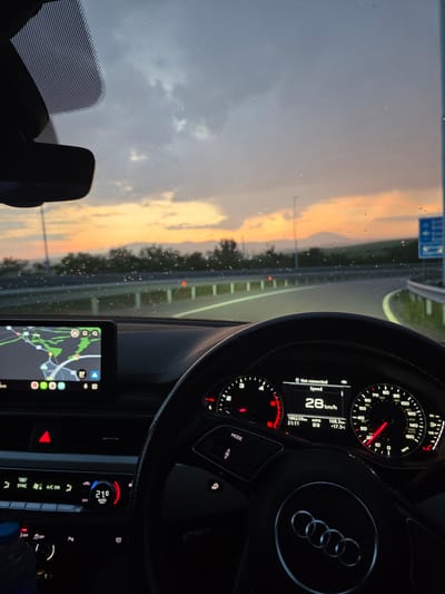 The image captures the interior of an Audi vehicle from the driver's perspective, looking out through the windshield at dusk in Balanovo, Bulgaria.
The vehicle's instrument cluster, featuring a black steering wheel with the Audi logo, displays "Not connected," a speed of 28 km/h, a total odometer reading of 186,239 km, a trip meter of 168.3 km, "AUTO" mode, gear selector D3, and an external temperature of +17.5°C. The time displayed is 21:11. The central infotainment screen shows a map application with green and grey areas, displaying a speed icon of "27 km/h" and several app icons at the bottom. Below the infotainment screen, the climate control panel indicates a setting of "21.0 AUTO," with the hazard light button illuminated red. Part of a blue bottle is visible on the lower left. Numerous small water droplets are present on the exterior of the windshield.
Beyond the windshield, a paved road, likely a highway, curves to the right. White lane markings are visible, and metal guardrails line the right side of the road. The sky transitions from an overcast grey overhead to vibrant orange, yellow, and red hues near the horizon, indicative of sunset or dusk. Distant hills or mountains are silhouetted against the colorful sky, with sparse green vegetation lining the road and terrain. A tall pole stands on the left side of the road, and a blue rectangular road sign with white arrows is partially visible on the right. No individuals are depicted in the scene.