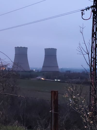 The image depicts an industrial landscape at twilight, centered on the Maritsa East 3 Thermal Power Plant in the vicinity of Mednikarovo, Bulgaria. Two prominent, grey cooling towers, illuminated with white lights around their upper rims, stand against an overcast, blue-grey sky. In the mid-ground, a winding road traverses green fields, with two vehicles visible by their illuminated headlights. Distant artificial lights of additional infrastructure are also present.

The foreground features natural and man-made elements. To the left, a dense cluster of bare tree branches and shrubs partially obstructs the view. A segment of a green wire fence is visible at the bottom edge. To the right, a rusty brown metal post stands vertically. Adjacent to it, a dark red metal lattice tower supports electrical lines and insulators, with two main power lines stretching horizontally across the upper sky. Bare tree branches with scattered white blossoms are also present in the lower right. No individuals are visible.
