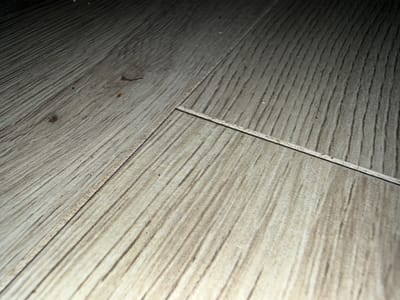 A close-up, low-angle photograph shows the surface of a light-colored laminate wood floor. The floorboards are laid parallel to each other, with distinct lines where the boards meet. The texture of the laminate mimics the grain of wood, with variations in light and dark brown striations running diagonally across the image. Some debris, appearing as small dark specks and a few orange-brown flecks, are visible on the floor's surface. The lighting suggests a dim environment, with a dark shadow cast across the upper portion of the image. The specific location in Hainstadt, Germany, is not discernible from the visual information provided.