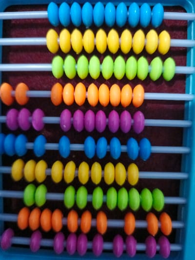 The image displays a colorful abacus, a traditional calculating tool, viewed from a slightly elevated, straight-on angle. The abacus features a light blue plastic frame, partially visible at the top, right, and bottom edges of the image. Within this frame, ten horizontal, light gray or white plastic rods are evenly spaced, running across a dark, reddish-brown, felt-like background.

Each rod holds ten brightly colored, oval-shaped plastic beads. The beads are arranged in a repeating color pattern for every five rows. From top to bottom, the color sequence is:
1.  **Top Row:** Blue beads. All ten blue beads are pushed to the left side of the rod.
2.  **Second Row:** Yellow beads. All ten yellow beads are pushed to the left side of the rod.
3.  **Third Row:** Lime green beads. All ten lime green beads are pushed to the left side of the rod.
4.  **Fourth Row:** Orange beads. Three orange beads are pushed to the left, and seven are on the right side of the rod.
5.  **Fifth Row:** Purple beads. Four purple beads are pushed to the left, and six are on the right side of the rod.
6.  **Sixth Row:** Blue beads. Five blue beads are pushed to the left, and five are on the right side of the rod.
7.  **Seventh Row:** Yellow beads. Six yellow beads are pushed to the left, and four are on the right side of the rod.
8.  **Eighth Row:** Lime green beads. Seven lime green beads are pushed to the left, and three are on the right side of the rod.
9.  **Ninth Row:** Orange beads. Eight orange beads are pushed to the left, and two are on the right side of the rod.
10. **Bottom Row:** Purple beads. Nine purple beads are pushed to the left, and one is on the right side of the rod.

The beads appear glossy, with some subtle reflections indicating a smooth surface. The lighting is bright and even, illuminating the vibrant colors of the beads. There are no people visible in the image, nor is there any visible text. The abacus seems to be clean and in good condition, suggesting it is either new or well-maintained. The arrangement of the beads in the lower rows indicates that various numbers are currently represented, consistent with the function of an abacus as a counting and calculation tool, possibly used for educational purposes or play.