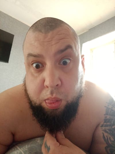 A close-up, unflattering portrait of a shirtless, bald, middle-aged man with a full black beard, positioned indoors with the top of his head close to the camera. His eyes are wide and appear slightly crossed, and he is sticking his tongue out slightly. The man's left hand is visible in the lower right corner, partially obscured by the beard, with a tattoo on his index finger. A larger tattoo is partially visible on his upper left arm. A television is partly visible on the wall to the left of the man. Light comes in from a window behind the man to the right. There is no indication of a location in the city of Chulman, Russia.